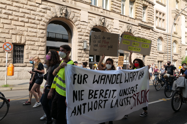 Eine Gruppe von Menschen marschiert bei einer Protestaktion gegen die Anti-Kriegs-Bewegung in Berlin, einige halten Schilder und andere fahren Fahrräder, im Hintergrund ein Gebäude mit Bögen, Säulen und Skulpturen.
