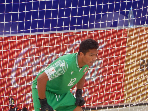 Ein Torwart in grüner Uniform kniet vor einem Tor mit Netz dahinter und einer Flasche rechts daneben.