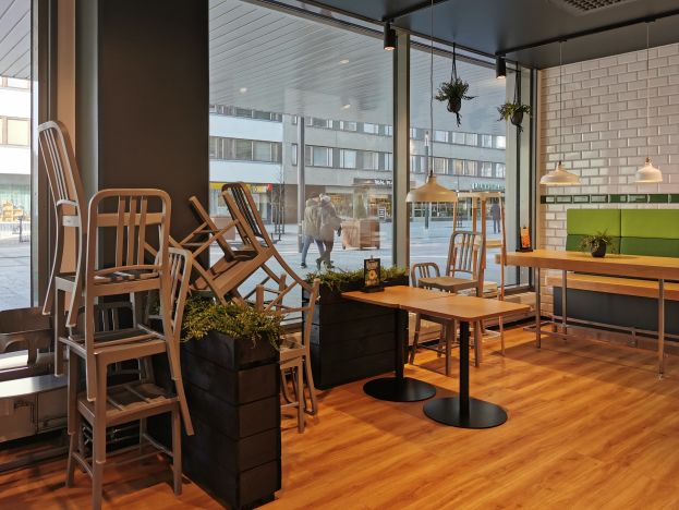 Ein Restaurantinnenraum mit Holztischen und -stühlen, großen Fenstern mit Blick auf eine belebte Straße in Berlin, Deutschland, umgeben von Pflanzen und Dekolights.