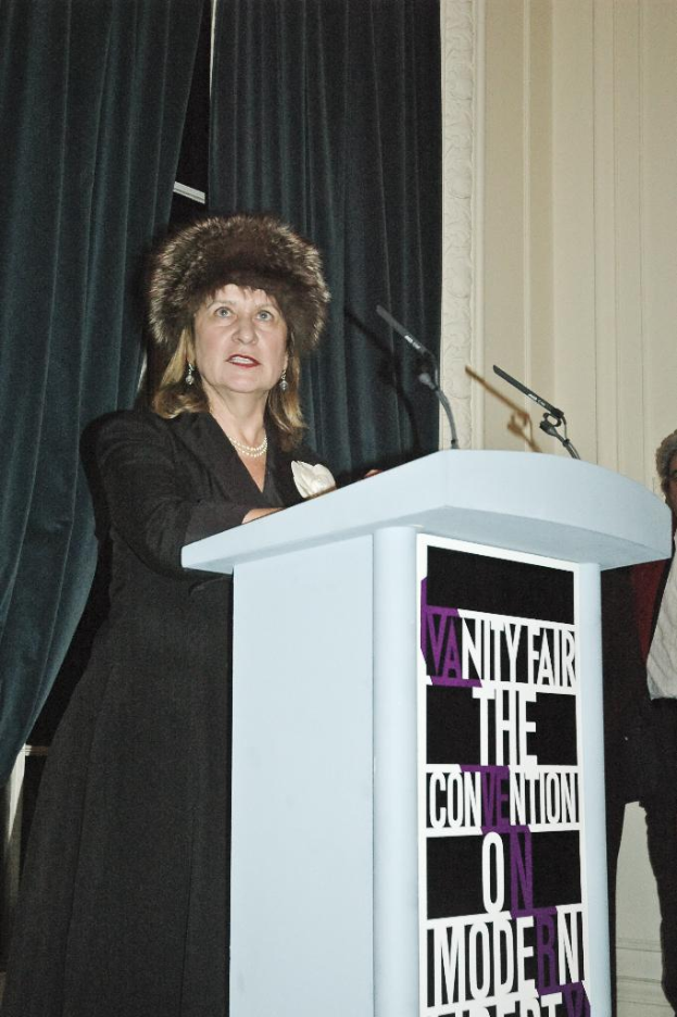 Eine Person steht neben einem Rednerpult und spricht in ein Mikrofon, mit mehreren Personen und Vorhängen im Hintergrund.