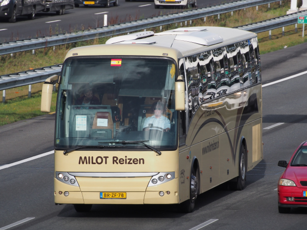 Ein Minibus mit sichtbaren Passagieren fährt auf einer Autobahn neben einem roten Auto, umgeben von Geländern und Gras, mit Textanzeigetafeln und Säulen im Hintergrund.
