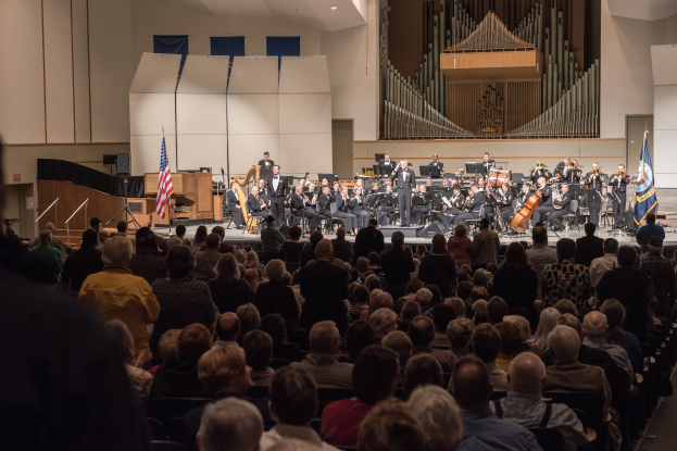Eine große Gruppe von Menschen sitzt vor einer B├╝hne mit einigen, die musikalische Instrumente halten. Flaggen mit St├Ąben, ein Lautsprecherkasten und andere Gegenst├Ąnde sind auf der B├╝hne zu sehen. An der R├╝ckwand befindet sich ein Dach mit Deckenleuchten und eine T├╝r, was auf ein Konzert an der University of Texas at Austin hindeutet.