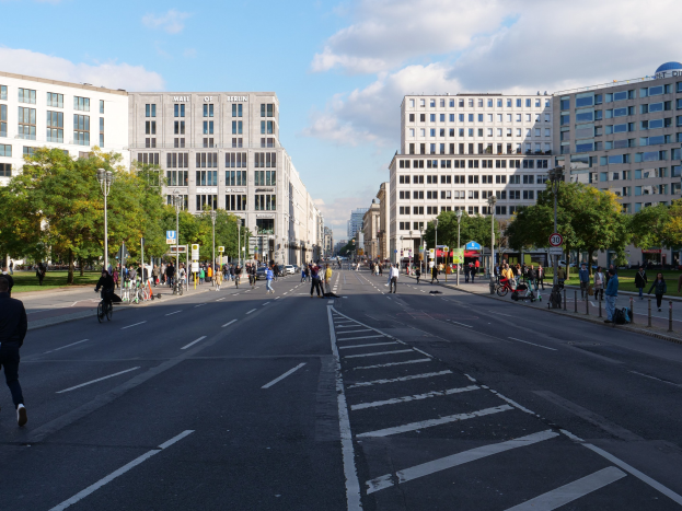 Eine belebte Stadtstraße in Berlin, Deutschland, mit Fußgängern und Radfahrern auf der Straße, hohen Gebäuden mit Fenstern, Bäumen, Laternen und Schildern unter einem bewölkten Himmel.