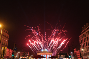 Eine belebte Stadtstraße am Silvesterabend in Berlin, voller Menschen, Fahrzeuge und Gebäude, erleuchtet von Lichtern und Feuerwerk am Himmel.