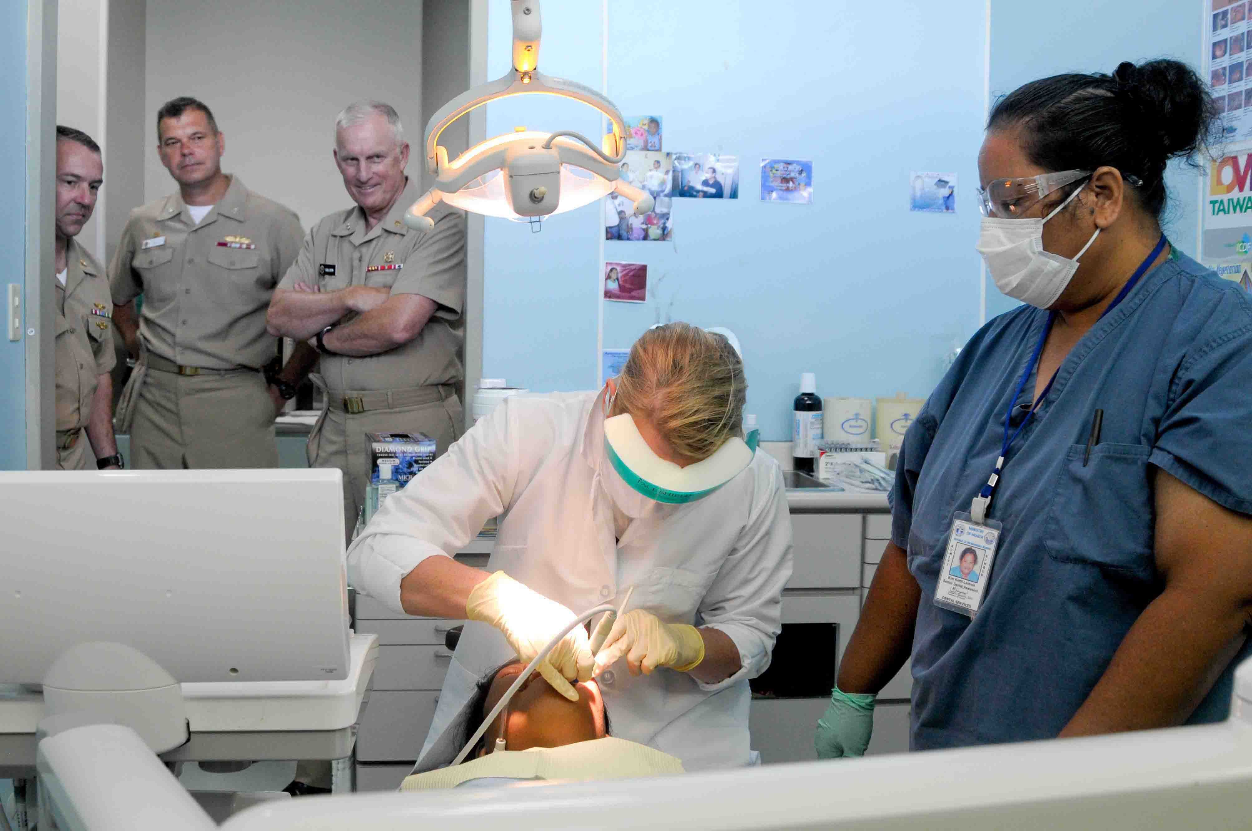 Eine Gruppe von Menschen in einer Zahnarztpraxis, mit einer Person in einem weißen Kleid, die ein Objekt hält, einer anderen mit Brille, Mundschutz und Ausweis, sowie Plakate an der Wand im Hintergrund.