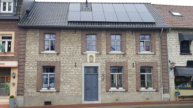 Ein Haus mit Solarpanelen auf dem Dach, Fenstern, Türen, Topfpflanzen, einer Bank, einem Schild, einem Straßenpfahl und einem bewölkten Himmel.