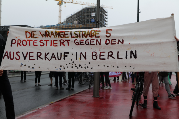 Eine Gruppe von Menschen steht auf der Straße und hält ein Schild hoch, auf dem "Die Wrangellstraße 5 protestiert gegen den Verkauf in Berlin" steht, mit Fahrrädern, Pfählen, Gebäuden und einem Kran im Hintergrund und einem sichtbaren Himmel.
