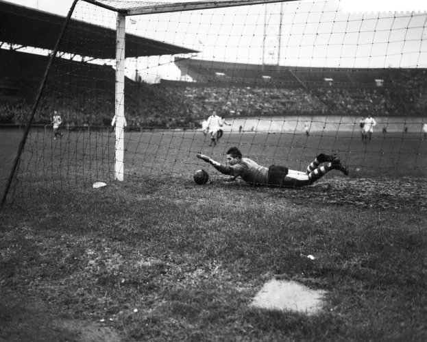 Ein Schwarz-Weiß-Foto eines Fußballspielers, der einen Ball vor einem Tor fängt, umgeben von anderen Spielern auf dem Boden und einem Stadion im Hintergrund.