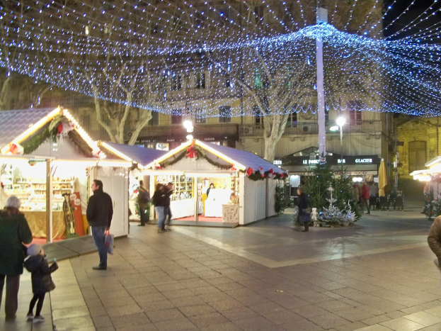 Ein lebendiger Weihnachtsmarkt in einer Stadtmitte bei Nacht, mit Menschen, die herumlaufen, beleuchteten Ständen und Gebäuden, Bäumen und Laternen im Hintergrund unter einem dunklen Himmel.