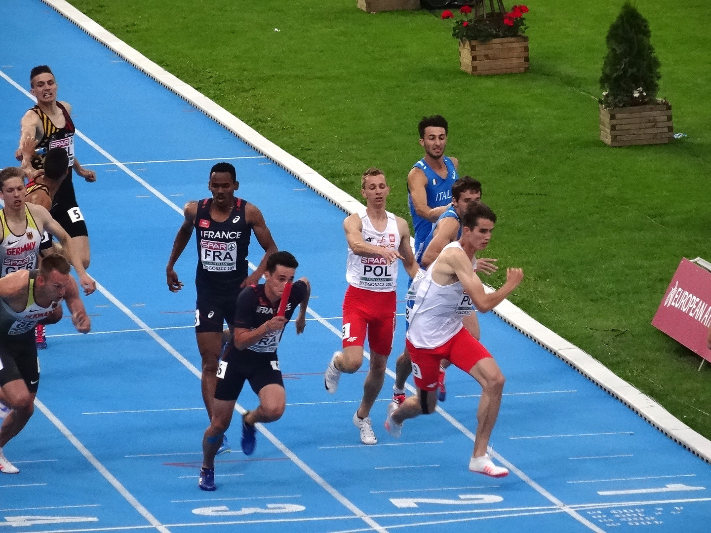 Eine Gruppe von Männern, die auf einer blauen Laufbahn umgeben von grünem Gras und Blumentöpfen laufen, mit einer Texttafel auf der rechten Seite, die auf einen Wettbewerb hinweist, wahrscheinlich die Europameisterschaften im Leichtathletik.