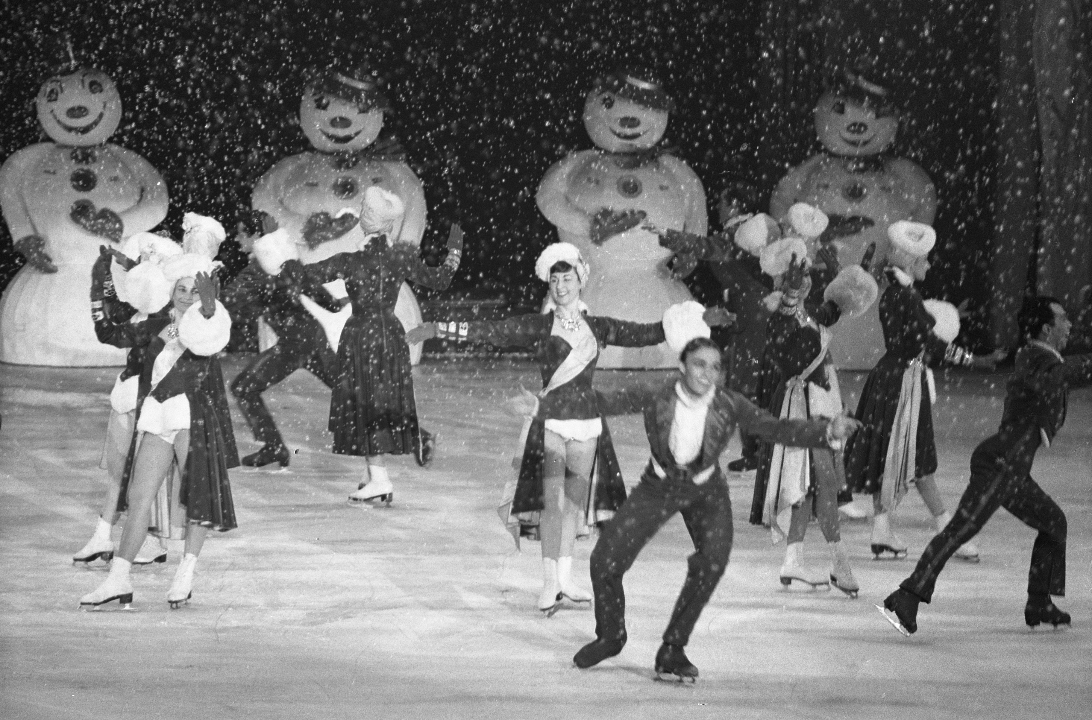 Eine Gruppe von Menschen in Kostümen und Schlittschuhschuhen ägyptisch auf einer Eisbahn im Schnee, mit Schneemännchen im Hintergrund, in Schwarz-Weiß aufgenommen.