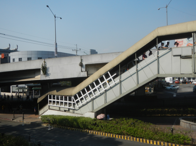 Eine Fußgängerbrücke mit Menschen, die über eine Straße gehen, umgeben von Gebäuden, Straßenlaternen, Strommasten, Stromkabeln, Kraftfahrzeugen, Büschen und einem klaren blauen Himmel.