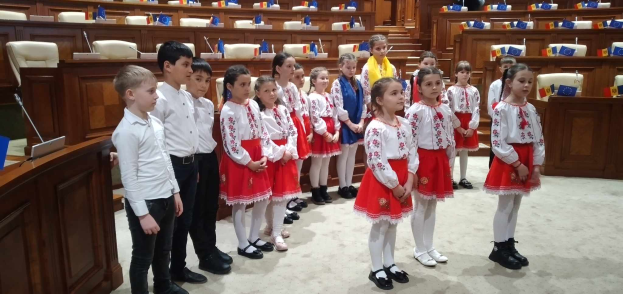 Eine Gruppe von Kindern in weißen und roten Kleidern steht zusammen in einem Raum mit einem Podium und einem Mikrofon links, Stühle und Tische im Hintergrund und Flaggen, die eine nationale Versammlung anzeigen.