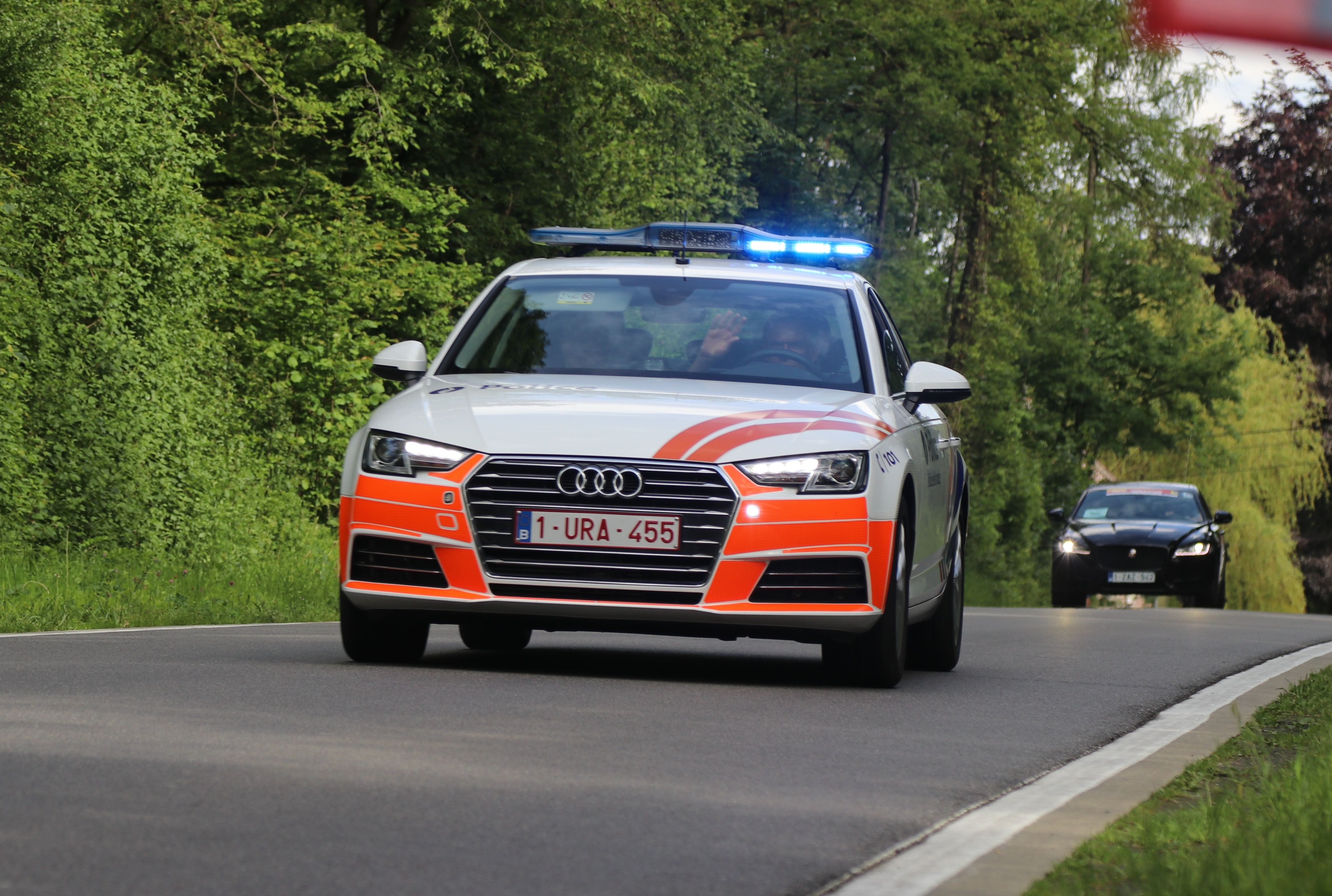Ein Audi A4 Polizeiwagen fährt auf einer Straße mit Grasrand, flankiert von einem Wald, mit zwei Insassen unter einem klaren blauen Himmel.