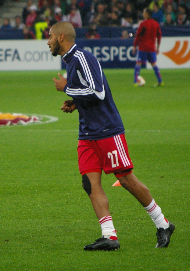 Ein Mann in einem blauen Hemd und roten Shorts läuft auf einem Fußballfeld, im Hintergrund sitzen einige Menschen und es sind ein paar Werbetafeln zu sehen.