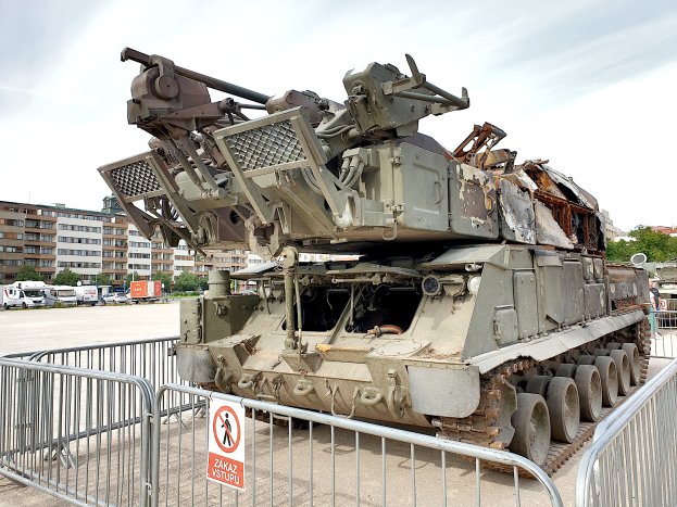 Ein gepanzertes Fahrzeug steht vor einem Metallzaun mit einem daran befestigten Schild, umgeben von Fahrzeugen, Bäumen, Gebäuden und einem klaren blauen Himmel.