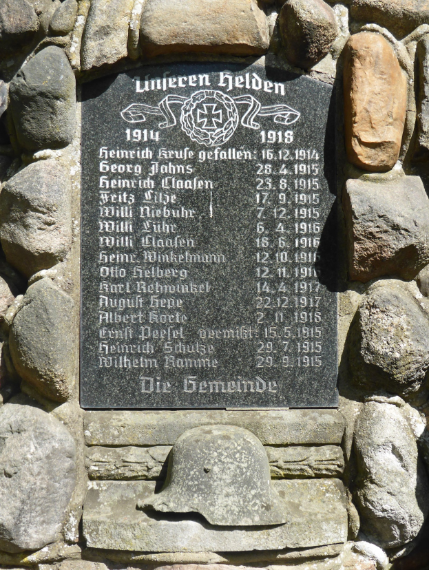 Steinmauer mit einer Gedenktafel und einem Helm darauf, beschriftet mit Text.