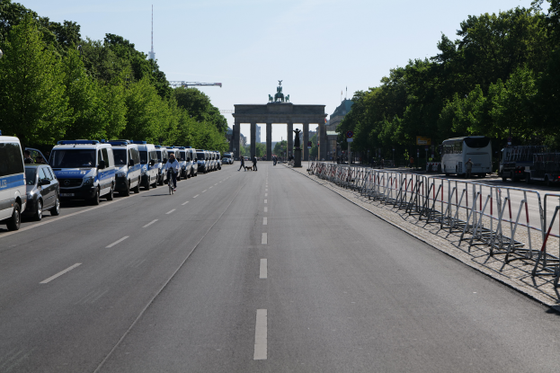 Eine Reihe von Polizeiwagen auf einer Straße vor dem Brandenburgertor in Berlin, Deutschland, mit Menschen auf Fahrrädern und Stehenden, Barrieren, Bäumen und einem Bogen mit Statuen im Hintergrund.