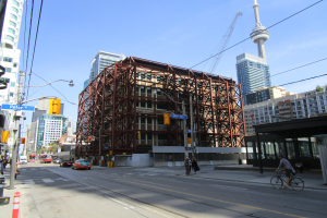 Eine belebte Stadtstraße mit einem großen Gebäude im Hintergrund, das gebaut wird, mit Fahrzeugen, einem Radfahrer, Fußgängern, Ampeln, Pfählen, Schildern, einem Kran und dem Himmel.