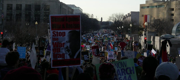 Eine Gruppe von Menschen, die auf einer Straße protestieren und Plakate halten, mit Gebäuden, Bäumen und Laternen im Hintergrund unter einem klaren Himmel.
