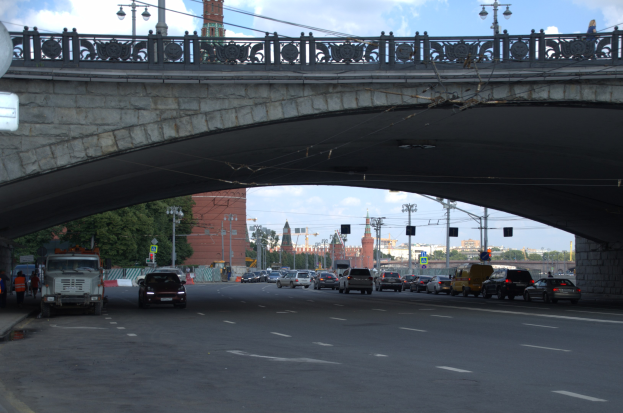Eine Brücke überspannt eine vielbefahrene Straße mit darunterfahrenden Fahrzeugen; Fußgänger stehen auf dem linken Gehweg, und im Hintergrund sind Laternenpfähle, Ampeln, Bäume, Gebäude und der Himmel zu sehen.