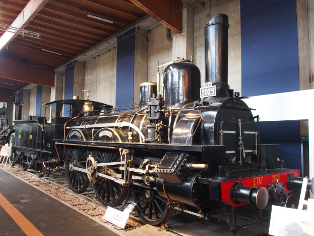 Eine alte Dampflokomotive in einem Museum ausgestellt, umgeben von Eisenbahnschienen, Informationsschildern, St├Ąnden und anderen Ausstellungsstücken.