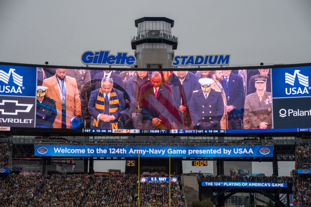 Eine große Menge Menschen in einem Stadion schaut ein Fußball-Spiel, mit einem zentralen Bildschirm, der Bilder und Text anzeigt, Schilder mit Text, Zuschauer in den Rängen, ein Tor, Bäume und einen klaren blauen Himmel im Hintergrund.