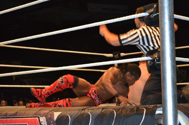 Zwei Männer kämpfen in einem Ring, einer trägt ein rotes Hemd und Schuhe, der andere Shorts, mit einem Schiedsrichter auf der rechten Seite und einer Zuschauermenge im Hintergrund unter einem dunklen Himmel.