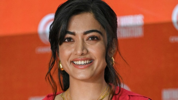 Eine Frau in einem roten Kleid mit einem eleganten Dutt und dezentem Make-up lächelt in die Kamera, trägt schwarze High Heels und hat ein gelbes Banner mit der Aufschrift "Sonam Kapoors beste Schönheits Looks vom Cannes Film Festival" im Hintergrund.