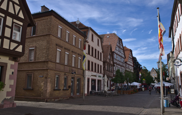 Headsteinstraße in Rothenburg ob der Tauber, Deutschland, gesäumt von Gebäuden mit Fenstern, Bäumen und Fahnenmasten, mit einigen Menschen und Fahrzeugen unter einem bewölkten Himmel.