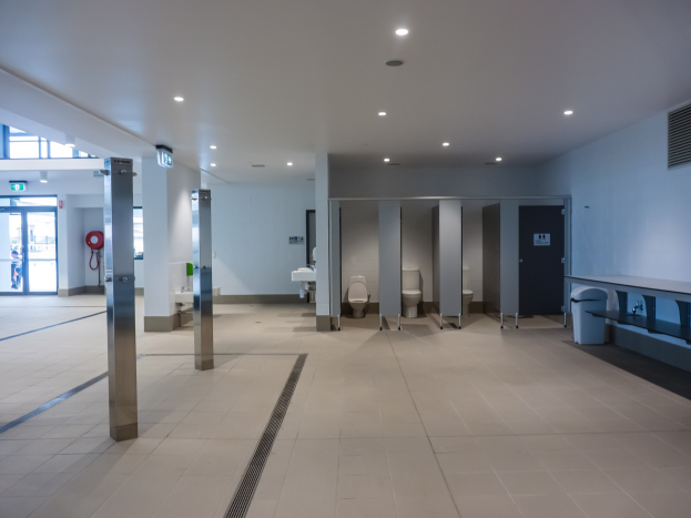 Große ßeffentliche Toilette mit mehreren Toiletten, tragenden Säulen, einem Mülleimer, einer Tür, einem Schild, einem Feuerlöscher und deckenmontierten Leuchten.