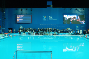 Eine Gruppe von Menschen, die um ein Hallenbad mit Netz am Boden stehen, mit einem Bildschirm im Hintergrund, der "FINA World Championships 2015 Kazan" anzeigt.