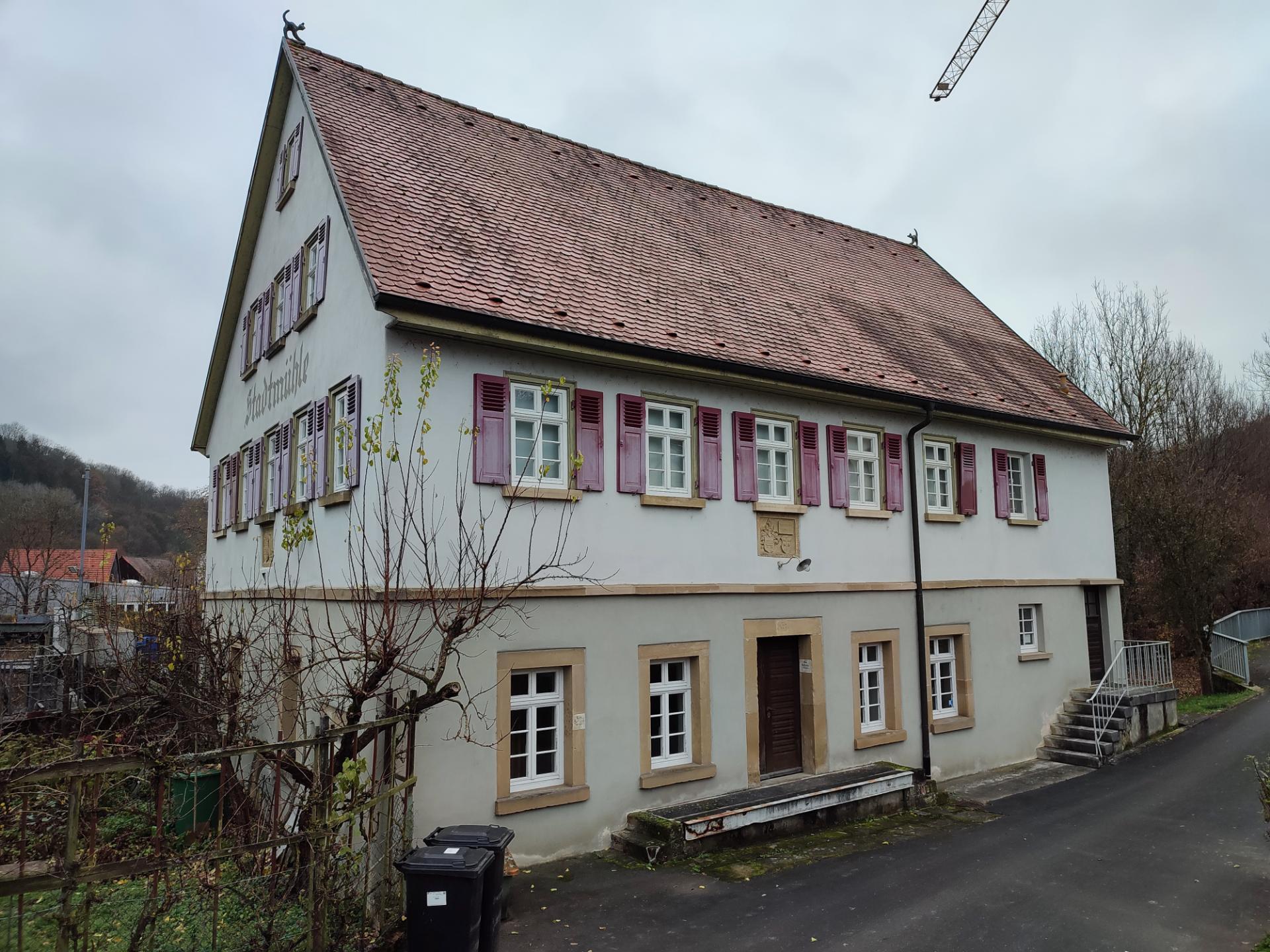 Ein großes weißes Gebäude mit roten Fensterläden, umgeben von Bäumen, Pflanzen und verschiedenen Gegenständen, mit Hügeln und einem klaren blauen Himmel im Hintergrund, zum Verkauf in Baden-Württemberg.