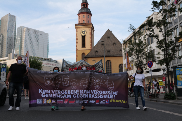 Eine Gruppe von Menschen mit Masken, die eine Fahne mit Text und Bildern halten, geht eine Straße entlang, mit einem geparkten Auto auf der linken Seite, Gebäuden, Bäumen, Schildern, Polen, einem Uhrenturm und einem klaren blauen Himmel im Hintergrund.