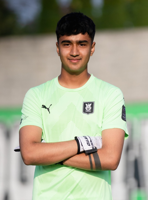 Ein junger Mann in einem grünen T-Shirt steht auf einem Fußballfeld und hat die Arme verschränkt, er lächelt, wahrscheinlich ein Torwart, mit einem unscharfen Hintergrund, der darauf hindeutet, dass der Fokus auf ihm liegt.