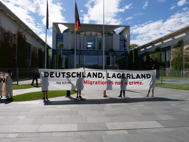 Gruppe von Menschen mit einem Transparent mit der Aufschrift 'Deutschland, Lagerland Migration ist kein Verbrechen' vor einem Gebäude mit Fenstern, umgeben von Gras, einem Metallzaun, Pflanzen, Bäumen und Fahnenstangen unter einem bewölkten Himmel.