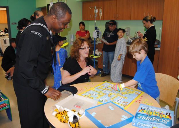 Gruppe von Menschen, die ein Brettspiel an einem Tisch mit Spielzeugen, Boxen und anderen Gegenständen spielen; Hintergrund enthält Schränke, Salzlösung auf einem Ständer, eine Tür und eine Wand.