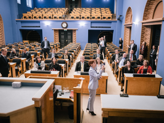 Frau hält Rede im schottischen Parlament, vor einem Publikum sitzend und stehend, mit Tischen mit Mikrofonen, Gläsern, Papieren und anderen Gegenständen, einer Uhr an der Wand und Deckenlampen im Hintergrund.