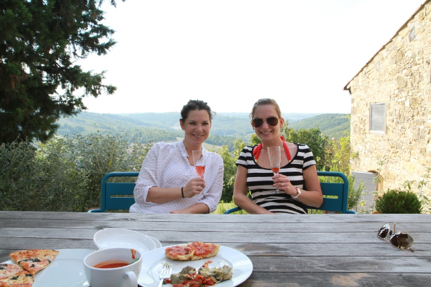 Zwei Frauen sitzen auf Stühlen im Freien, lächeln und halten Gläser in der Hand, mit einem Tisch dazwischen, der Goggles, eine Tasse, Pizza-Scheiben auf einem Teller und eine Gabel zeigt.