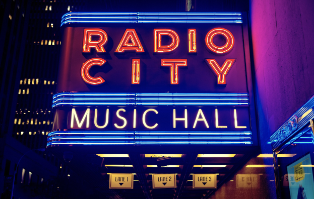 Außenansicht des Radio City Music Hall in New York City bei Nacht, hell erleuchtet mit hohen Gebäuden im Hintergrund, mit einem "Radio City"-Schild und zusätzlichen Schildern auf der rechten Seite.