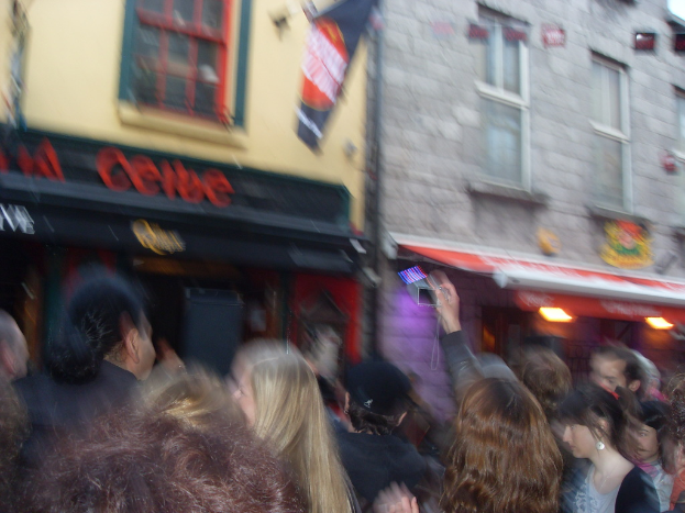 Ein unscharfer Bildausschnitt, der Menschen auf der Straße unten und Gebäude mit Lichtern, Fenstern und einer Fahne oben zeigt.