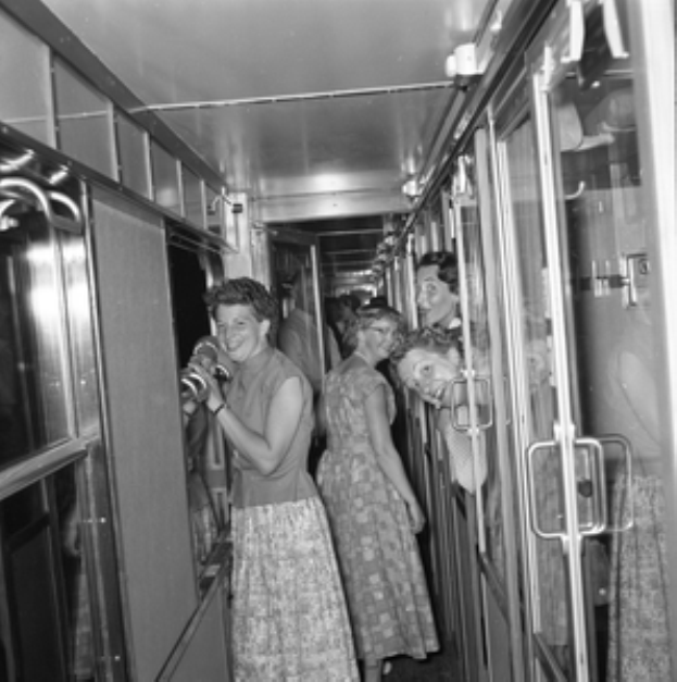Eine Gruppe von Frauen, die auf einem Zug mit Türen auf beiden Seiten und Lichtern öber ihnen steht, dargestellt in Schwarz-Weiß.