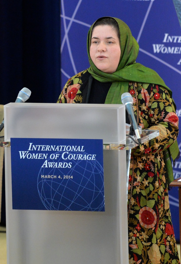 Eine Frau steht an einem Podium mit einem Mikrofon, einer Tafel am Podium und einem Tisch rechts daneben, mit einem Banner im Hintergrund, auf dem 'International Women of Courage Awards 2014' geschrieben steht.