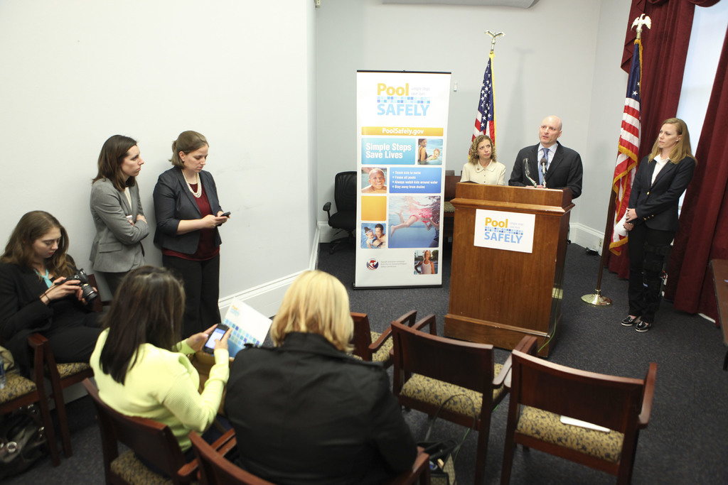 Drei sitzende Frauen, ein Mann und eine Frau vor einem Rednerpult, zwei Fahnen und ein Banner im Hintergrund, eine Frau mit einer digitalen Kamera auf der linken Seite, zwei Frauen neben ihr stehend und Vorhänge im oberen rechten Eck.