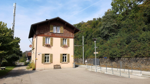 Ferienhaus zum Verkauf in Baden-Württemberg, Deutschland, mit einem mehrfenstrigen Gebäude hinter einer Hecke, umgeben von Straßen, Grünflächen und einem bewölkten Himmel.