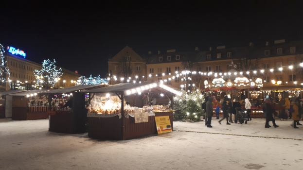 Weihnachtsmarkt mit Menschen, Buden, Pflanzen und Gebäuden unter einem bewölkten Nachthimmel in einer verschneiten Straße.