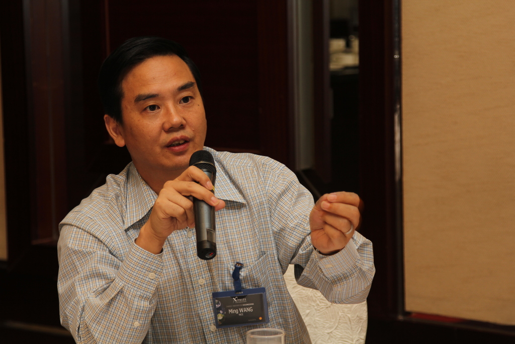 Ein Mann in einem Hemd hält ein Mikrofon und spricht, mit einem Abzeichen auf seiner Tasche, vor einer Wand im Hintergrund.