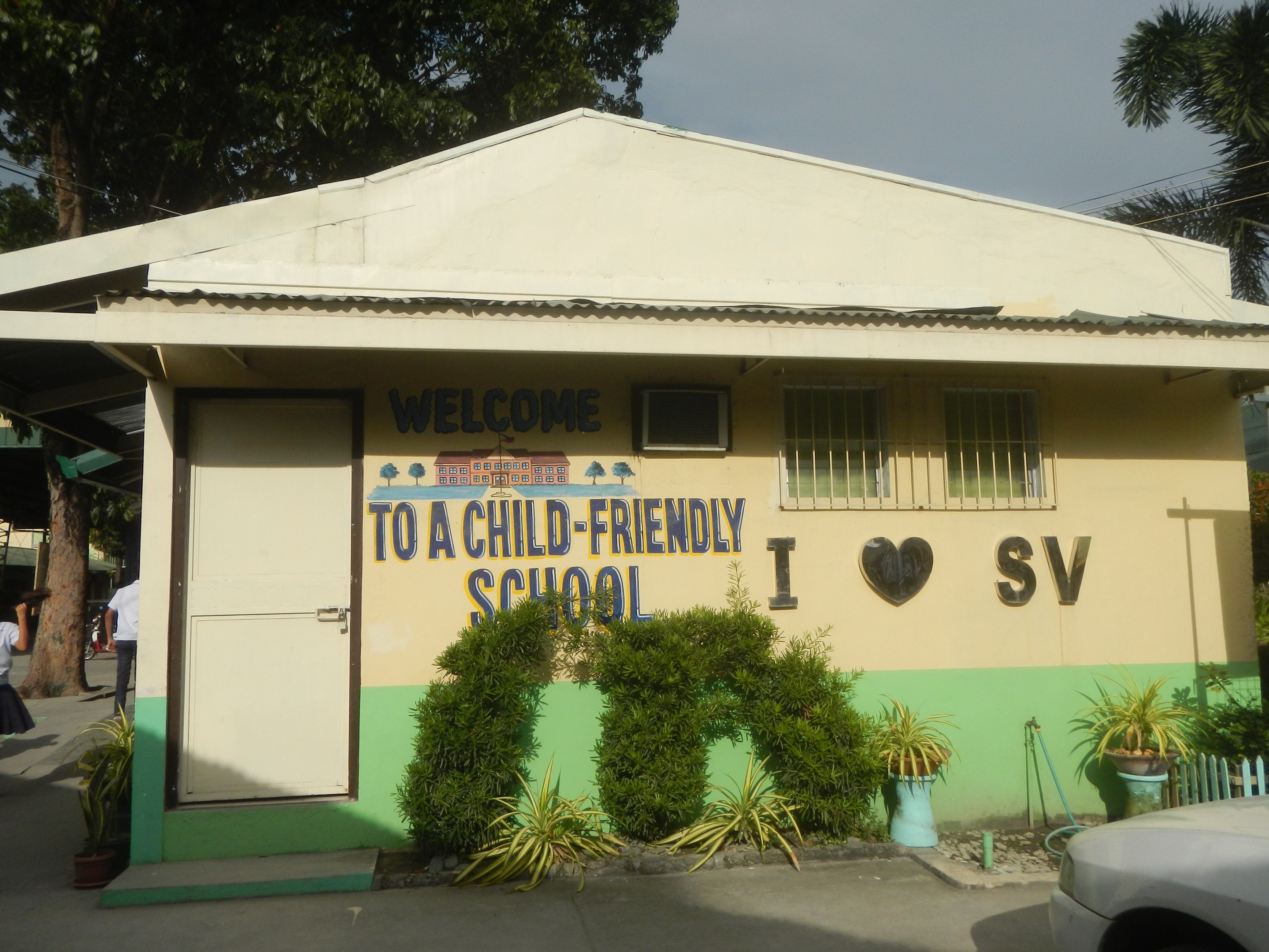Ein einstöckiges Schulhaus mit einem davor geparkten Auto, das eine Tür, Fenster und ein "Willkommen in einer kinderfreundlichen Schule"-Schild sowie umliegende Topfpflanzen, Bäume und Menschen mit sichtbaren Strommasten, Drähten und Baumrinde unter einem bewölkten Himmel zeigt.