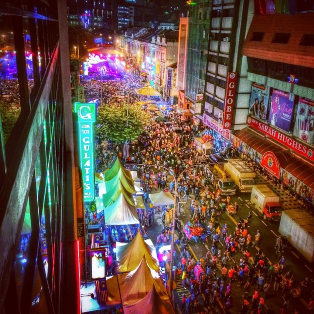 Volles Karnevalsbild mit Zelten, Buden, Gebäuden und Bannern, mit beleuchteter Fläche im Hintergrund.