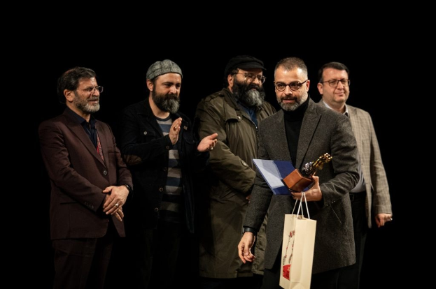 Männer in festlicher Kleidung auf einer Bühne, einer hält eine Tasche und eine Trophäe, mit einem dunklen Hintergrund, der eine Feier an der Eröffnungsnacht der Berliner Filmfestspiele andeutet.